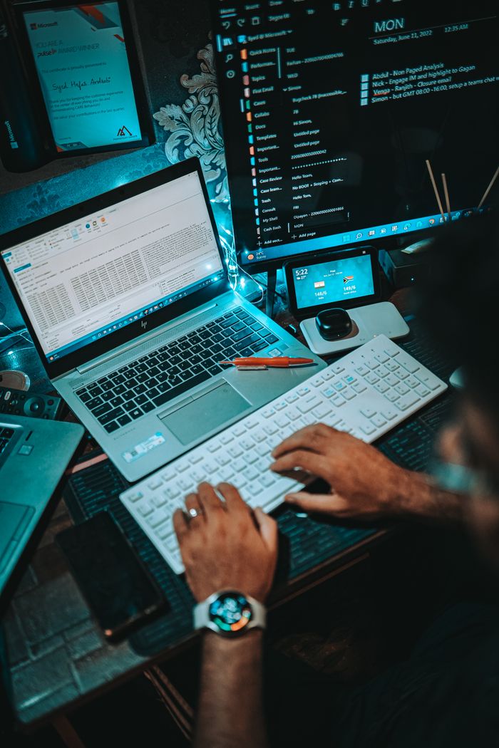 Services A person working on a dual-monitor setup with a laptop and keyboard in an office environment.